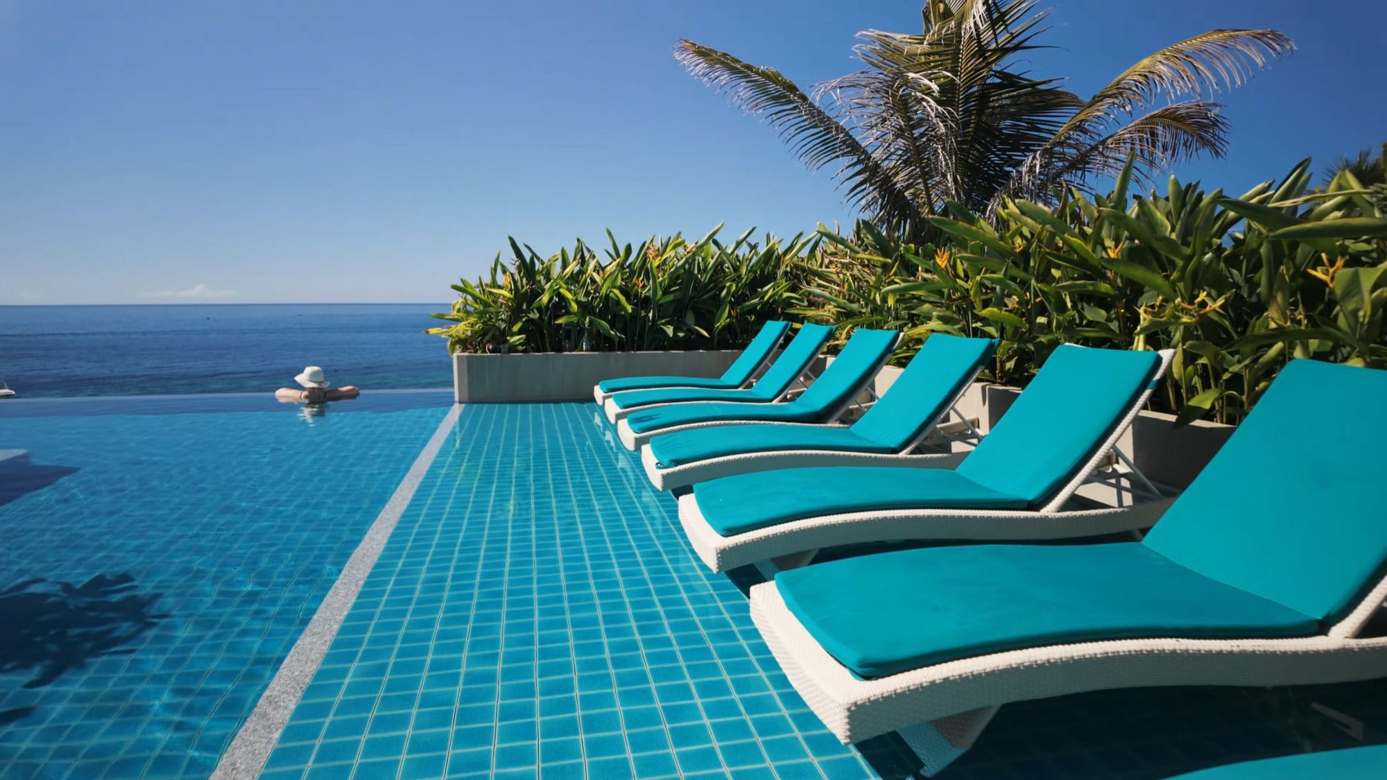 A row of turquoise sun loungers beside an infinity pool, the sea beyond, one swimmer in a straw hat at the edge