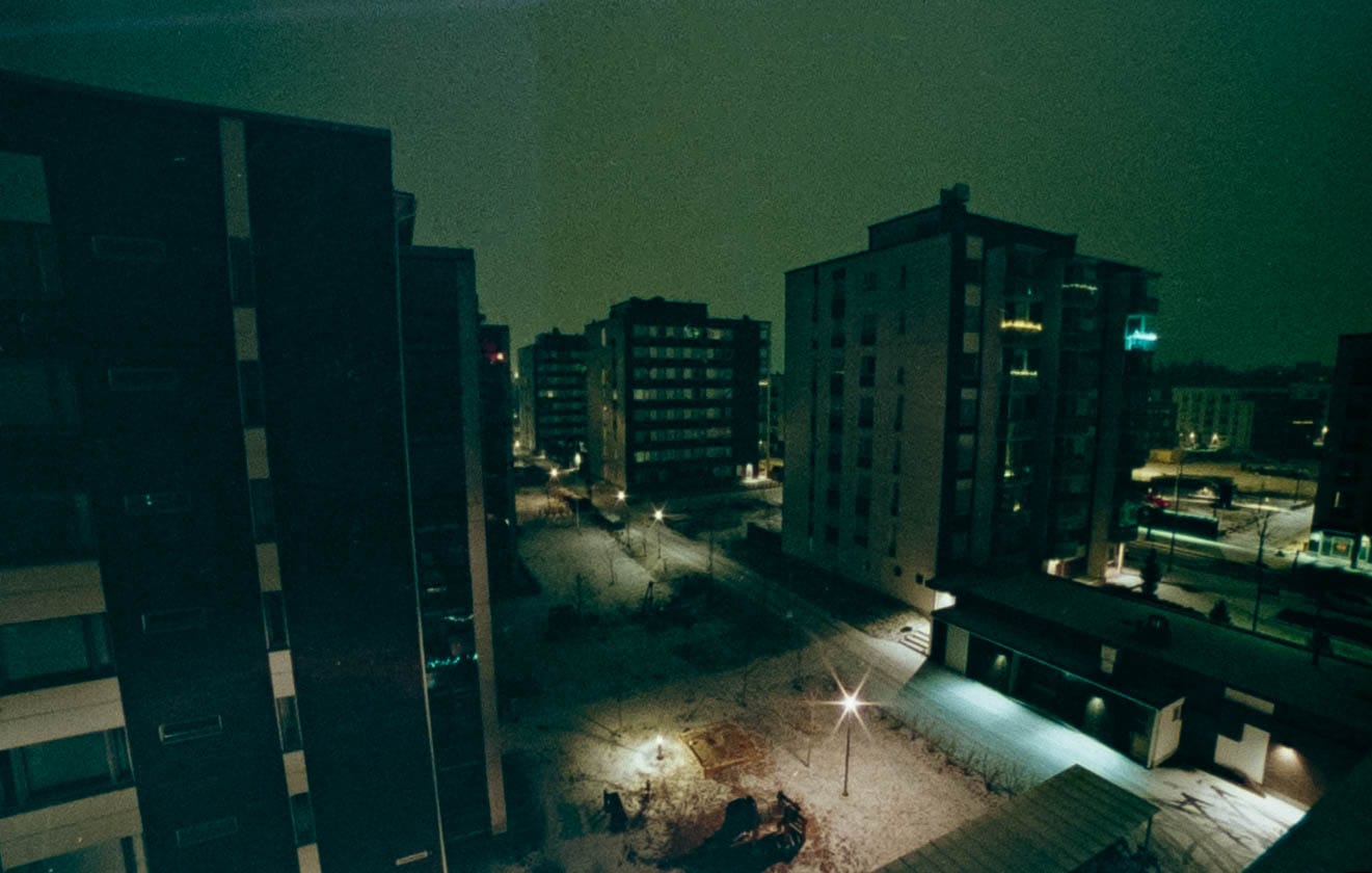 Apartment blocks around a snow-dusted courtyard at night, lit by a few street lamps, on film