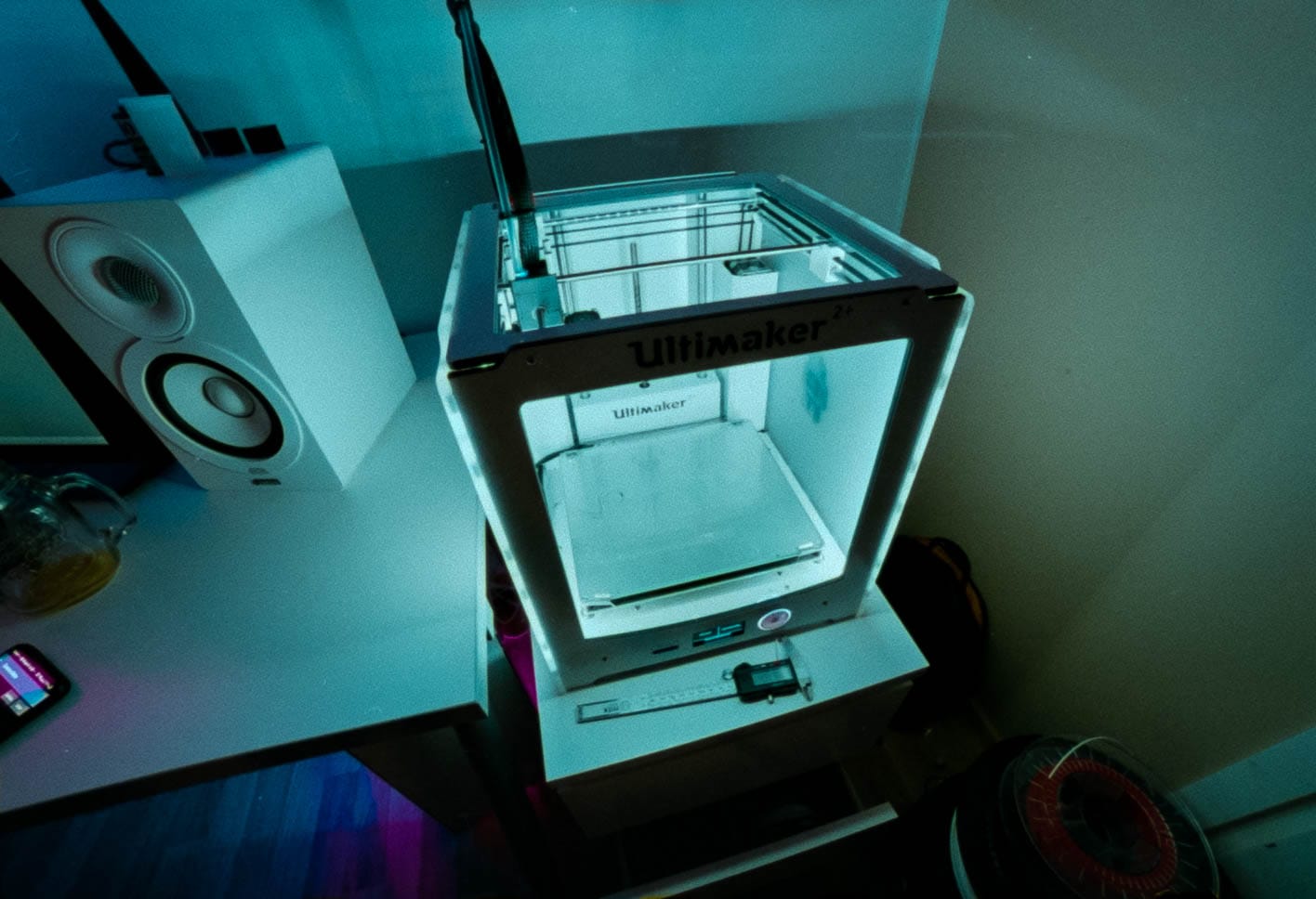 An Ultimaker 2+ printer glowing on a desk beside a speaker, on film