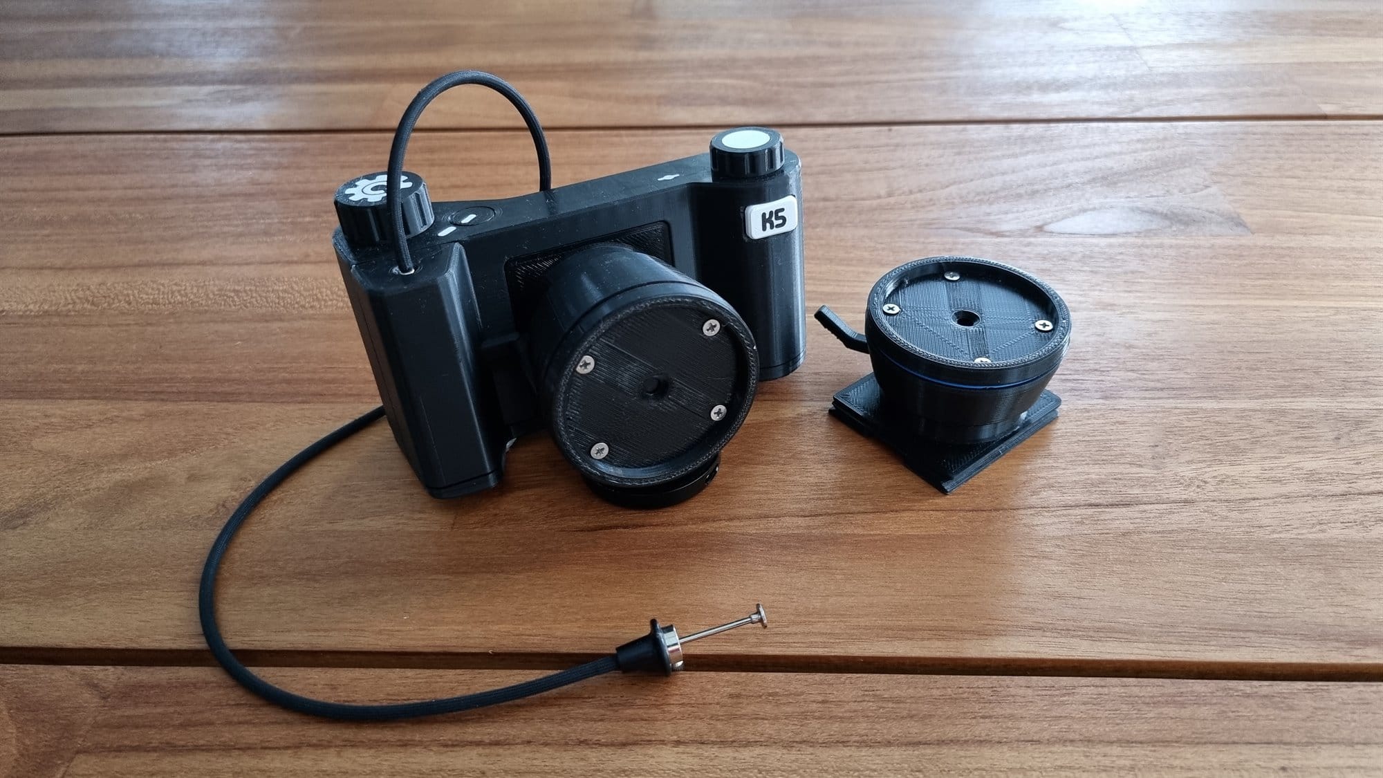 The black 3D-printed camera on a wooden table with a cable release attached and a second lens unit beside it